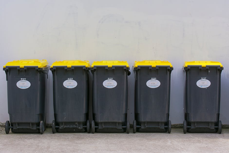 A row of five large wheeled rubbish bins made of black plastic with textured surfaces, positioned on a concrete pavement against a light grey wall. Each bin has a hinged yellow lid with a slightly rai