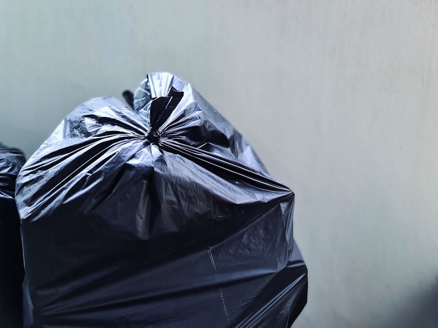 Close-up of a tightly secured black plastic rubbish bag with a glossy finish, resting against a plain, light-colored wall. The bag shows creases, folds, and slight crumpling, indicating it is filled w
